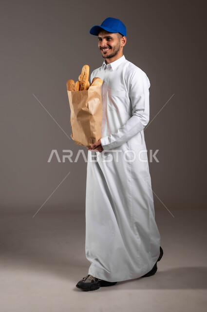 الانطلاق لاداء العمل بهمة ونشاط، شراء المواد الغذائية المختلفة من خلال الانترنت، المخبوزات والمعجنات الطازجة الشهية، خدمة التوصيل السريع في السعودية، بورتريه لمندوب عربي خليجي سعودي  يرتدي الزي التقليدي و قبعه زرقاء، صورة لكامل الجسم، خلفيه رماديه