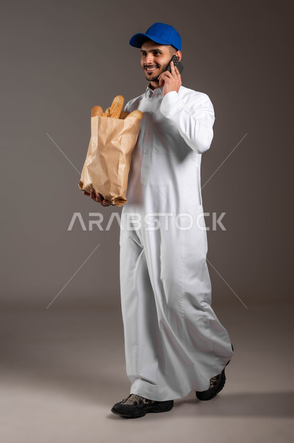 التواصل مع الزبائن عبر الموبايل، الاستفادة من التكنولوجيا الحديثة بتسهيل العمل، المخبوزات والمعجنات الطازجة الشهية، خدمة التوصيل السريع في السعودية، بورتريه لمندوب عربي خليجي سعودي  يرتدي الزي التقليدي و قبعه زرقاء، صورة لكامل الجسم، خلفيه رماديه