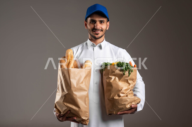 ايماءات الرضا لانجاد العمل، الخضروات والمخبوزات الطازجة، شراء الاحتياجات المنزلية والمواد الغذائية المختلفة من خلال الانترنت، بورتريه مقرب لمندوب توصيل عربي خليجي سعودي  يرتدي الزي التقليدي وقبعه زرقاء يقوم بتسلم اكياس ورقيه، خلفيه رماديه