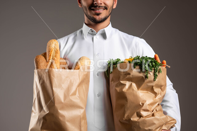 Fresh vegetables and baked goods, express delivery service in Saudi Arabia, purchasing household needs and various food items through the Internet, a close-up portrait of a Saudi Gulf Arab representative wearing traditional clothing receiving paper bags, gray background