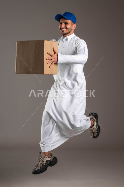 إيماءات السعادة والنشاط، بورتريه من الجانب لمندوب عربي خليجي سعودي مبتسم يرتدي الزي التقليدي و القبعة الزرقاء يحمل بده صندوق، خدمة التوصيل السريعة والآمن في السعودية، عملية نقل الطلبات للزبائن، صورة لكامل الجسم، خلفيه رماديه