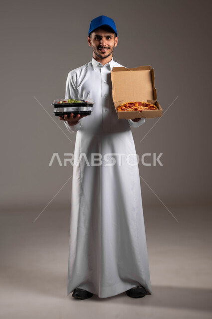 شراء الاحتياجات والمواد الغذائية المختلفة من خلال خدمة الديليفري، بورتريه لمندوب توصيل عربي خليجي سعودي  يرتدي الزي التقليدي وقبعه زرقاء يحمل بيده علبة بيتزا وسلطة خضار، تسليم الوجبات بأمان للزبائن، الأطعمة والمعجنات الطازجة، صورة لكامل الجسم، خلفيه رماديه