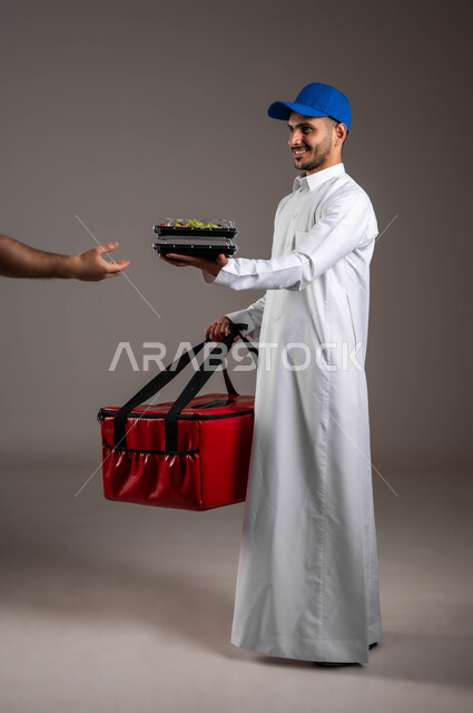 ايماءات السرور والسعاده لوصول الطلبيه بشكل آمن، بورتريه  لشاب عربي خليجي سعودي مبتسم يرتدي الزي التقليدي يضع قبعه زرقاء يمسك بيده شنته حمراء، طلبات الدليفري لوجبات الطعام الصحيه، صوره لكامل الجسم،  خلفيه ملونه