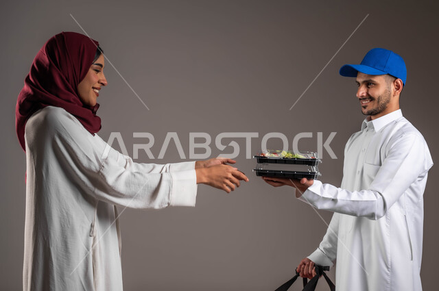 خدمات التوصيل السريع، بورتريه مقرب لصبيه عربيه خليجيه سعوديه ترتدي العباءه البيضاء والحجاب الاحمر تقوم باستلام المشتريات من مندوب التوصيل، حمل طبق من السلطه الخضراء الشهيه, طلبات الدليفري لوجبات الطعام الصحيه، خلفيه رماديه