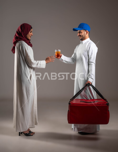 ايماءات السعادة باستلام كأس العصير من المندوب بشكل سليم، بورتريه من الجانب لشاب عربي خليجي سعودي مبتسم يرتدي الزي التقليدي  وقبعة زرقاء يحمل بيده شنتة حمراء، عملية شحن وتوصيل موثوقه وناجحة، صوره الجسم بالطول الكامل، خلفية رماديه
