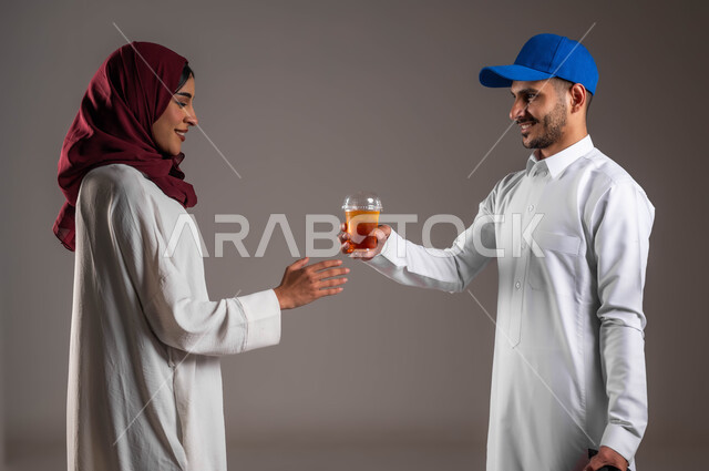 الاستمتاع بتناول العصائر البارده، بورتريه مقرب من الجانب لرجل عربي خليجي سعودي مبتسم يرتدي الزي التقليدي وقبعة زرقاء يحمل بيده كوب عصير، خدمة توصيل المشروبات الى المنزل، ارتداء العباءة البيضاء والحجاب الأحمر، خلفية رماديه