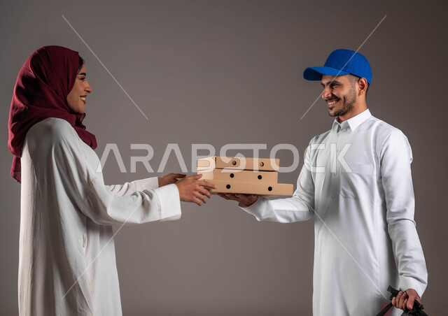 خدمات التوصيل السريع الآمن، بورتريه لفتاه عربية خليجية سعودية مبتسمة ترتدي العباءه البيضاء والحجاب الاحمر تقوم باستلام كراتين الطعام من المندوب، ايماءات السعاده لوصول الطلبيه بشكل سليم، ارتداء الزي التقليدي والقبعة الزرقاء، خلفيه رماديه