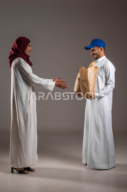 التسوق وشراء الطعام الصحي من خلال خدمة الديليفري، بورتريه من الجانب لشاب عربي خليجي سعودي يرتدي الملابس التقليديه ويضع قبعة زرقاء يمسك بيده اكياس ورقيه من المخبوزات والخضار، ارتداء عباءة بيضاء وحجاب أحمر، صوره الجسم بالطول الكامل، خلفيه رماديه