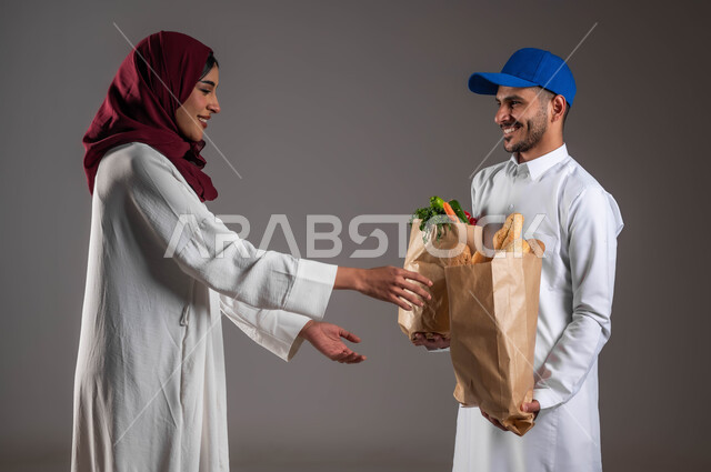 شراء الاحتياجات المنزلية والمواد الغذائية المختلفة من خلال خدمة الديليفري، بورتريه من الجانب لمندوب توصيل عربي خليجي سعودي  يرتدي الزي التقليدي وقبعه زرقاء يسلم اكياس ورقيه من الخضروات والمخبوزات للعميلة، ارتداء عباءة بيضاء وحجاب أحمر، خلفيه رماديه