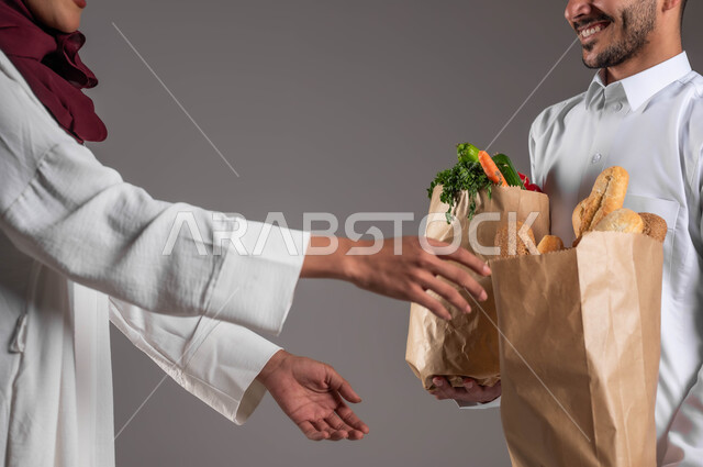 الحفاظ على الوزن المثالي من خلال تناول الخضراوات، بورتريه مقرب من الجانب لامرأه عربيه خليجيه سعوديه ترتدي عباءه بيضاء وحجاب احمر تستلم المشتريات من مندوب التوصيل، أكياس ورقية تحتوي على المخبوزات والخضار، خلفيه رمادية