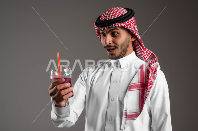 تزويد الجسم بالطاقة من خلال تناول المشروبات الصحيه، بورتريه مقرب لشاب عربي خليجي سعودي يرتدي الملابس التقليديه والشماغ وينظر الى كوب العصير بتعابير الصدمة والاستغراب، توفير العناصر الغذائية الضرورية للصحة، ايماءات الدهشة بلذة المشروب الطازج، خلفيه رماديه
