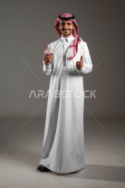 تحسين المزاج وإعطاء شعور بالانتعاش، بورتريه لرجل عربي خليجي سعودي مبتسم يرتدي الشماغ والملابس التقليديه يشير بإصبع الإبهام بعلامة القبول والتأكيد، ايماءات الاعجاب بطعم المشروب الصيفي المنعش، صورة الجسم بالطول الكامل، خلفيه ملونه