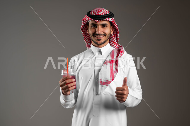 إيماءات السعادة والاستمتاع بتناول المشروبات الباردة، بورتريه مقرب لشاب عربي خليجي سعودي يرتدي الشماغ والملابس التقليديه يشير بإبهامه بعلامة الاعجاب، محاولة التخفيف من حرارة الصيف بالمشروب المنعش، خلفيه رمادية