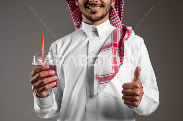 ايماءات السعادة والاعجاب بطعم المشروب الطازج، بورتريه لرجل عربي خليجي سعودي يرتدي الثوب التقليدي والشماغ يحمل بيده كوب عصير ويشير بعلامة الجودة والامتياز، مشروبات صيفية منعشة ومثلجة، خلفية رماديه