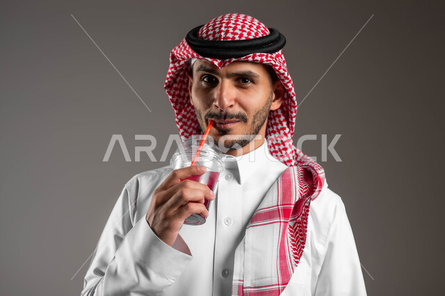 تحسين المزاج واعطاء شعور بالانتعاش، بورتريه مقرب لرجل عربي سعودي خليجي مبتسم يرتدي الشماغ والملابس التقليديه ويمسك بيده كوب من العصير، محاولة التخفيف من حرارة الصيف بالمشروب الطبيعي، خلفية رماديه