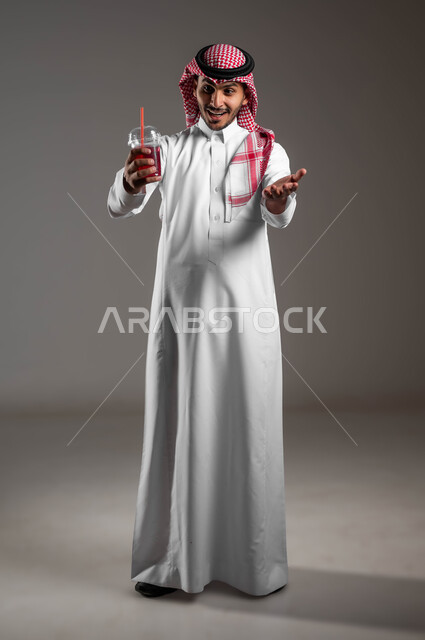 تزويد الجسم بالطاقة من خلال تناول المشروبات الصحيه، بورتريه لشاب عربي خليجي سعودي مبتسم يرتدي الزي التقليدي والشماغ يحمل بيده كوب من العصير الطبيعي، مشروب صيفي منعش ومثلج، تحسين المزاج واعطاء شعور بالانتعاش، صوره لكامل الجسم، خلفية رماديه