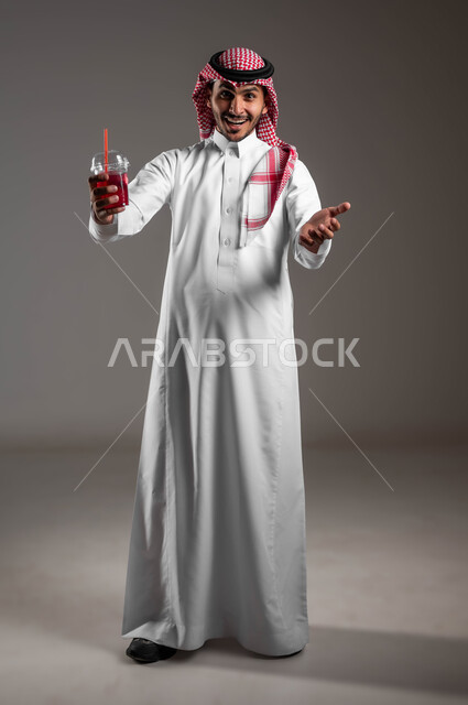 ايماءات السعادة والسرور اثناء تناول المشروبات الباردة، بورتريه لشاب عربي خليجي سعودي يرتدي الزي التقليدي والشماغ يمسك بيده كوب من العصير الطبيعي، تحسين مستويات الطاقة والتركيز خلال اليوم، صورة الجسم بالطول الكامل، خلفيه رماديه