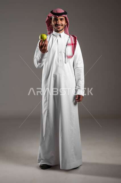 وجبات دايت صحية خفيفة، محاصيل زراعية سعودية ذات جودة عالية، تحسين مستويات الطاقة والتركيز خلال اليوم من خلال تناول الفواكه، بورتريه  لرجل عربي سعودي خليجي يرتدي الملابس التقليديه والشماغ يحمل بيده حبة من التفاح اللذيذ، صوره الجسم بالطول الكامل، خلفيه رماديه