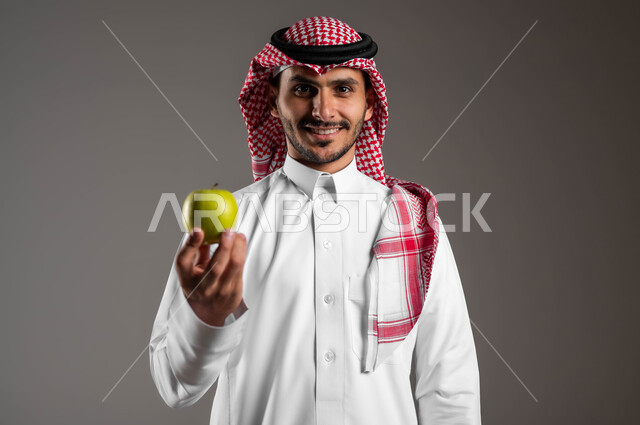 تلبية احيتاجات الجسم من العناصر الغذائية الضرورية, بورتريه مقرب لرجل عربي سعودي خليجي مبتسم يرتدي الملابس التقليديه والشماغ يحمل بيده حبة تفاح، الحفاظ على الوزن المثالي وتجنب المأكولات الضارة، محاصيل زراعية سعودية ذات جودة عالية، خلفيه رمادية