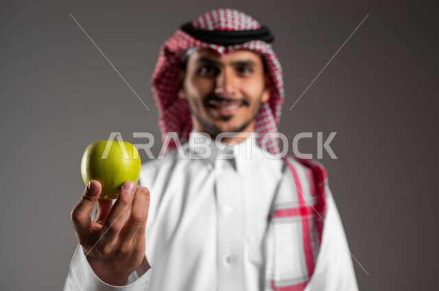انتقاء أجود أنواع الفواكه، محاصيل زراعية سعودية ذات جودة عالية، بورتريه مقرب لرجل عربي سعودي خليجي مبتسم يرتدي الملابس التقليديه والشماغ يحمل بيده تفاحه، تلبية احتياجات الجسم من الفيتامينات، تحسين اللياقة البدنيه من خلال الطعام الصحي، خلفيه رماديه