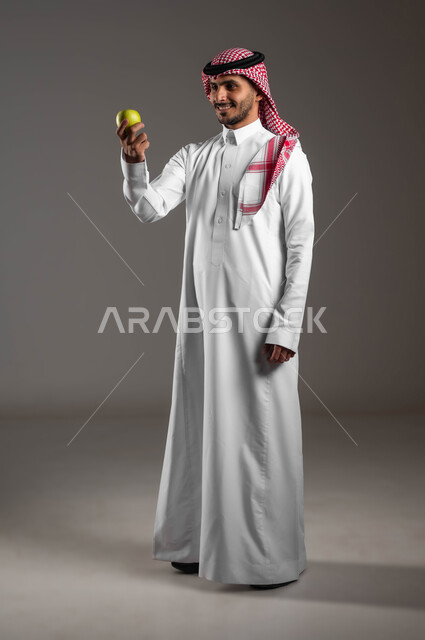 الحفاظ على صحة الجسم وتقليل خطر الاصابة بالأمراض، محاصيل زراعية سعودية ذات جودة عالية، بورتريه  لرجل عربي سعودي خليجي مبتسم يرتدي الملابس التقليديه والشماغ ويحمل بيده حبة تفاح، انتقاء أفضل أنواع الفواكه، تجنب المأكولات الضارة، صوره الجسم بالطول الكامل، خلفيه رماديه
