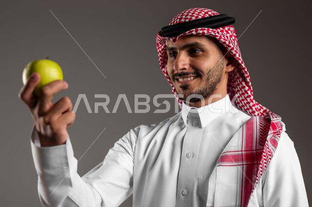 الحفاظ على الوزن المثالي وتجنب المأكولات الضارة، محاصيل زراعية سعودية ذات جودة عالية، بورتريه مقرب لشاب عربي سعودي خليجي مبتسم يرتدي الملابس التقليديه والشماغ يحمل بيده فاكهة التفاح اللذيذة، توفير العناصر الغذائية الضرورية للجسم، خلفية رماديه