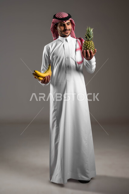 نظام غذائي صحي معتمد على الموز والاناناس، محاصيل زراعية سعودية ذات جودة عالية،  بورتريه لرجل عربي خليجي سعودي يرتدي الزي التقليدي والشماغ ويمسك بيديه حبات الفاكهة الشهية، الحفاظ على الوزن المثالي وتجنب المأكولات الضارة، صوره الجسم بالطول الكامل، خلفيه رمادية