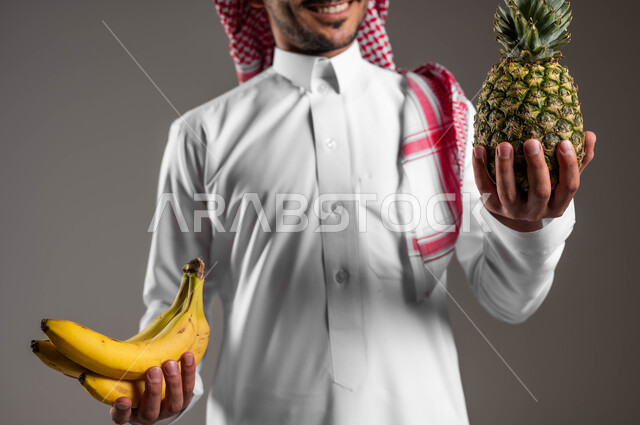 الحفاظ على الرشاقة والصحه البدنية خلال اليوم من خلال تناول الفواكه، بورتريه مقرب لشاب عربي خليجي سعودي يرتدي الزي التقليدي والشماغ يمسك بيديه حبات الموز والاناناس، محاصيل زراعية سعودية ذات جودة عالية، تلبية احتياجات الجسم من الفيتامينات، خلفية رماديه