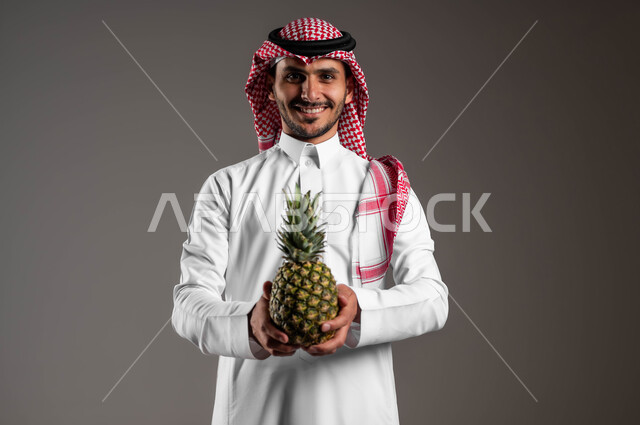 تلبية الحاجات الضرورية للجسم، انتقاء أجود أنواع الفواكه الطازجة، بورتريه لشاب عربي خليجي سعودي يرتدي الزي التقليدي والشماغ مبتسم ينظر الى الكاميرا ويحمل بيده حبة أناناس، المحاصيل الزراعية السعودية ذات الجودة العالية، وجبة طعام صحية خفيفة، خلفية رمادية