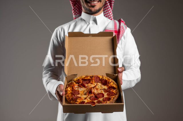 ضرر تناول وجبات طعام غير صحية، طلبات الأطعمة الخارجية، أصناف المعجنات والفطائر الشهية، بورتريه مقرب لرجل عربي خليجي سعودي يرتدي الثوب التقليدي والشماغ يحمل البيتزا في علبة كرتون، مطاعم المأكولات الغربية في السعودية، خلفية رمادية