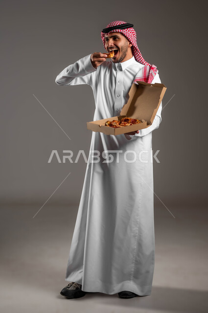 Receiving food through delivery orders, delicious fresh and hot pastries, portrait of a Saudi Gulf Arab man wearing a traditional dress and shemagh eating delicious pizza, not counting the calories consumed, eating an unhealthy meal, full length of the body, gray background