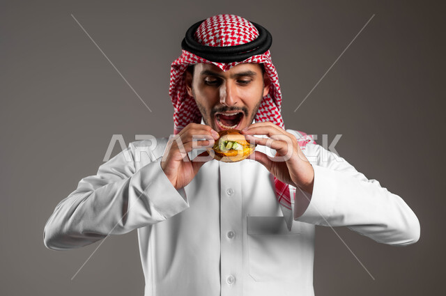 اضرار الإكثار من أكل الوجبات السريعة، استلام وطلب الأطعمة الخارجية، بورتريه مقرب لشاب عربي خليجي سعودي يرتدي الثوب التقليدي والشماغ يقوم بتذوق ساندويش البرجر اللذيذ، تناول الطعام الغير الصحي، مطاعم للمأكولات الغربية في السعودية، خلفية رمادية