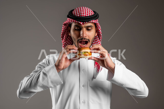 استلام وطلب الأطعمة الخارجية، ضرر الإكثار من أكل الوجبات السريعة، بورتريه مقرب لشاب عربي خليجي سعودي يرتدي الثوب التقليدي والشماغ يقوم بتذوق ساندويش البرجر اللذيذ، تناول الطعام الغير الصحي، مطاعم للمأكولات الغربية في السعودية، خلفية رمادية
