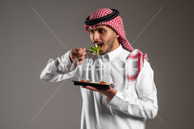 توفير العناصر الغذائية الضرورية للجسم، تناول وجبات الطعام الصحية، بورتريه مقرب لشاب عربي خليجي سعودي يرتدي زي تقليدي وشماغ يقوم بتذوق طبق من السلطة الشهية، الحفاظ على الرشاقة والصحة البدنية والتقليل من خطر الإصابة بالأمراض، خلفية رمادية