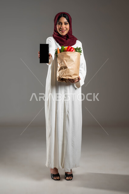 كيس ورقي يحتوي على الخضراوات الطازجة، مفهوم التسوق الالكتروني , شراء الأطعمه من السوبرماركت عبر الاونلاين,  بورتريه لسيدة عربية خليجية سعودية مبتسمة ترتدي العباءة البيضاء والحجاب الأحمر وتمسك الهاتف المحمول بشاشة سوداء فارغة، صورة الجسم بالطول الكامل، خلفية رمادية