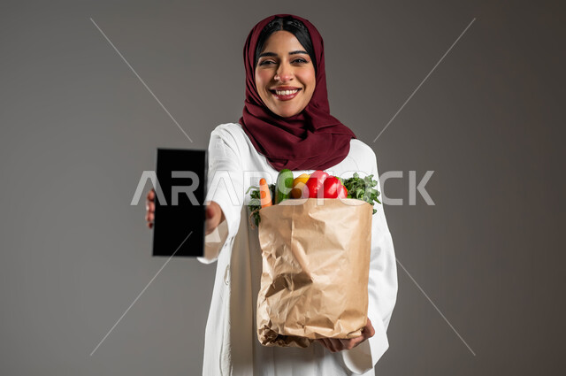 مفهوم التسوق الالكتروني , شراء الأطعمه من السوبرماركت عبر الاونلاين,  بورتريه مقرب لسيدة عربية خليجية سعودية مبتسمة ترتدي العباءة البيضاء والحجاب الأحمر وتمسك الهاتف المحمول بشاشة سوداء فارغة، كيس ورقي يحتوي على الخضراوات الطازجة، خلفية رمادية