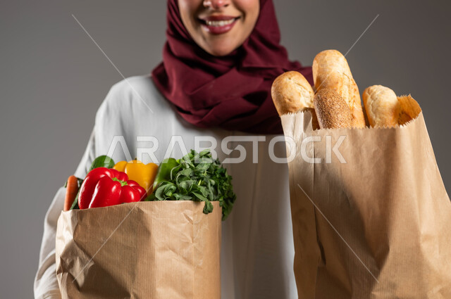 شراء الخبز والخضراوات من المتجر، بورتريه مقرب لسيدة عربية خليجية سعودية مبتسمة ترتدي العباءة البيضاء والحجاب الأحمر تحمل بيدها أكياس ورقية من المأكولات الصحية، الحفاظ على الوزن المثالي من خلال تناول الطعام الصحي، خلفية رمادية