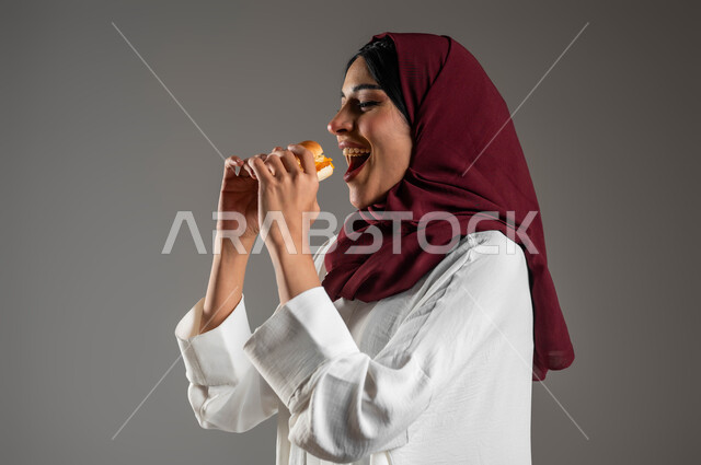 الوجبات السريعة والشهية،  مفهوم الطعام الغير صحي، ايماءات الاستمتاع بتناول الطعام، توفير الوقت والجهد في التحضير والطهي، بورتريه مقرب من الجانب لامرأه عربية خليجية سعودية ترتدي العباءة البيضاء والحجاب الأحمر وتتناول البرجر، خلفية رمادية