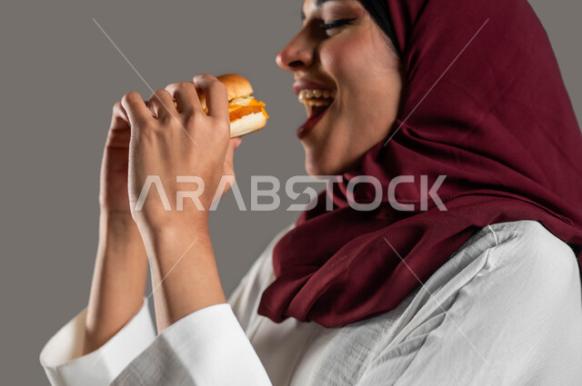 ايماءات الاستمتاع بتناول الساندويش، مفهوم الطعام الغير صحي،  توفير الوقت والجهد في التحضير والطهي، بورتريه مقرب من الجانب لشابة عربية خليجية سعودية ترتدي العباءة البيضاء والحجاب الأحمر وتتناول البرجر، وجبات الطعام السريعة الشهية، خلفية رمادية