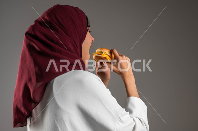 توفير الوقت والجهد في التحضير والطهي، بورتريه مقرب من الخلف لسيدة عربية خليجية سعودية ترتدي العباءة البيضاء والحجاب الأحمر وتتناول البرجر، مفهوم الطعام الغير صحي،  وجبات الطعام السريعة الشهية، خلفية رمادية