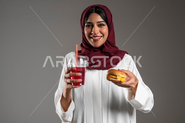 تناول المشروبات الباردة الشهية، مفهوم الطعام الغير صحي،  بورتريه مقرب لفتاة عربية خليجية سعودية ترتدي العباءة البيضاء والحجاب الاحمر وتحمل بيدها ساندويش البرجر اللذيذ وكأس من العصير، ايماءات السعادة والاستمتاع بالوجبة السريعة، خلفية رمادية