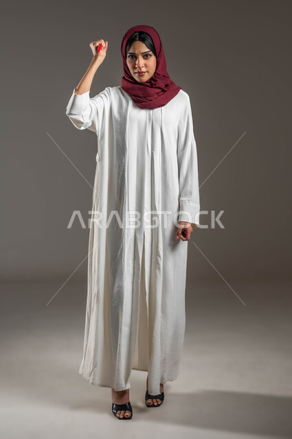 المشاكل الزوجية والعلاقات الاسريه الفاشلة، الامراض النفسيه والمشاكل الزوجيه , بورتريه لشابة عربية خليجية سعودية ترتدي عباءه بيضاء مع حجاب احمر تحمل بيدها سكين وتنظر الى الكاميرا بغضب، القتل للدفاع عن النفس ضد العنف الأسري، صورة الجسم بالطول الكامل، خلفية رمادية