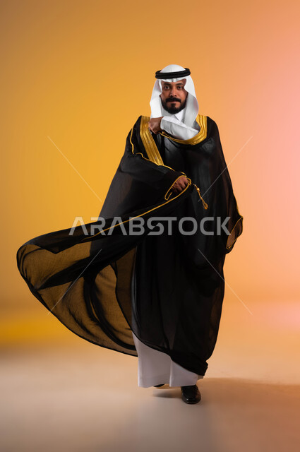 أناقة وفخامة اللباس الرسمي التقليدي  للمناسبات الخاصة والأعراس، السرور والفرح في ليلة الزفاف،  بورتريه لعريس عربي خليجي سعودي يرتدي البشت الاسود والغترة، ايماءات الحب والسعادة لبدء حياة جديدة، صورة لكامل الجسم، خلفية ملونة
