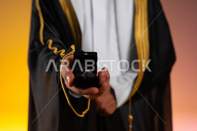 العادات والتقاليد للتقدم والطلب الزواج، شراء مجوهرات ومحبس العروس، مفهوم الاستقرار وتكوين عائلة سعيدة، بورتريه مقرب لعريس عربي خليجي سعودي يرتدي الغترة التقليدية والبشت الاسود ويمسك بيده خاتم الزواج، مراسم الأعراس السعودية، خلفية ملونة
