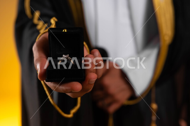 شراء مجوهرات ومحبس العروس، العادات والتقاليد للتقدم والطلب الزواج، مفهوم الاستقرار وتكوين عائلة سعيدة، بورتريه مقرب لعريس عربي خليجي سعودي يرتدي الغترة التقليدية والبشت الاسود ويمسك بيده خاتم الزواج، مراسم الأعراس السعودية، خلفية ملونة