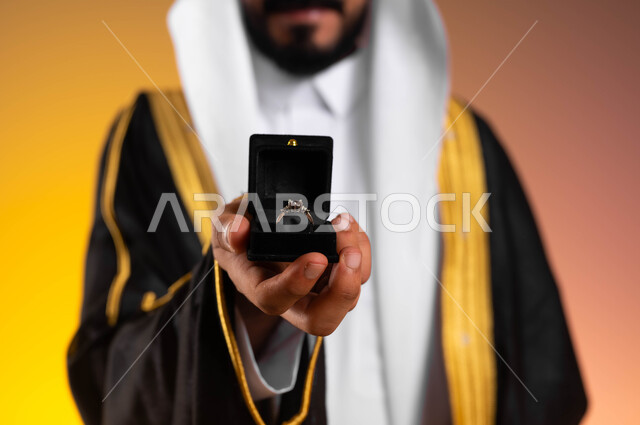 مفهوم الاستقرار وتكوين عائلة سعيدة، بورتريه مقرب لعريس عربي خليجي سعودي يرتدي الغترة التقليدية والبشت الاسود ويمسك بيده خاتم الزواج، مراسم الأعراس السعودية، العادات والتقاليد للتقدم والطلب الزواج، شراء مجوهرات ومحبس العروس، خلفية ملونة