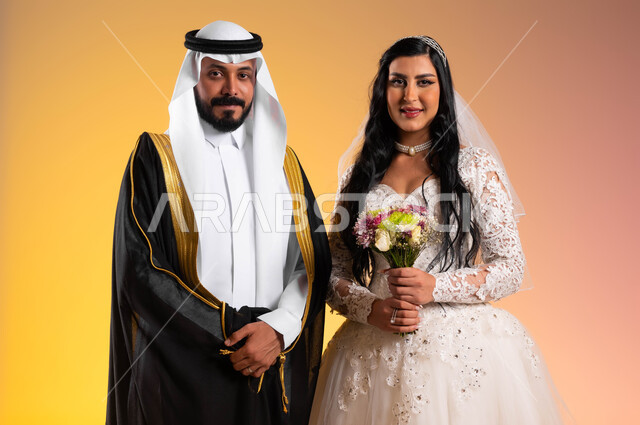 بدء حياة جديدة، النظر الى الكاميرا بتعابير السرور والفرح، مفهوم الزواج والاستقرار وتكوين عائلة، أصالة عادات وتقاليد مراسم الزواج في السعودية، بورتريه مقرب لعروس عربية خليجية سعودية مبتسمة ترتدي فستان الزفاف الابيض تحمل باقة الورود تقف بجانب زوجها، خلفية ملونة