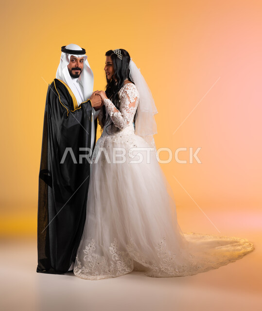 نظرات مليئة بالرومنسية والحب، مراسم الأعراس والحفلات في المملكة، مفهوم التزيين والاطلالة المتأنقة، بورتريه لعروس عربية خليجية سعودية مبتسمة ترتدي فستان الزفاف الأبيض تنظر الى زوجها بتعابير المودة, صورة الجسم بالكامل، خلفية ملونة