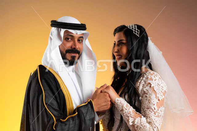مفهوم الزواج والاستقرار وتكوين عائلة، نظرات مليئة بالرومنسية والحب، مراسم الأعراس والحفلات في المملكة، مفهوم التزيين والاطلالة المتأنقة، بورتريه مقرب لعروس عربية خليجية سعودية مبتسمة ترتدي فستان الزفاف الأبيض تمسك يد زوجها بتعابير المودة, خلفية ملونة