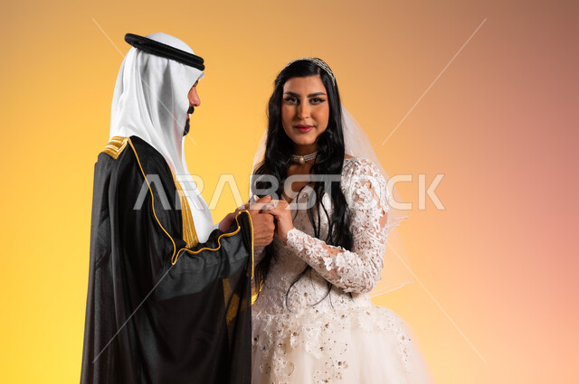 مفهوم الزواج والاستقرار وتكوين عائلة، نظرات مليئة بالرومنسية والحب، مراسم الأعراس والحفلات في المملكة، مفهوم التزيين والاطلالة المتأنقة، بورتريه مقرب لعروس عربية خليجية سعودية مبتسمة ترتدي فستان الزفاف الأبيض تمسك يد زوجها بتعابير المودة, خلفية ملونة